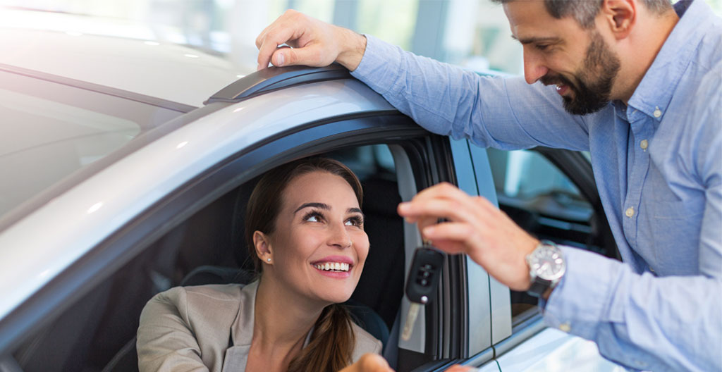 Conseillère automobile professionnelle remettant les clés d'un véhicule électrique compact neuf blanc à un jeune couple souriant devant un concessionnaire automobile moderne français